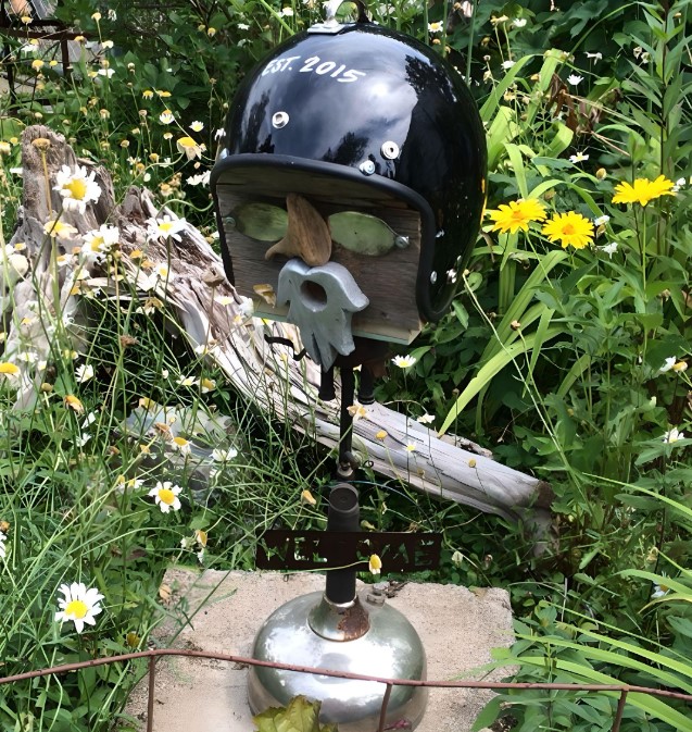 An upcycled birdhouse fashioned from a vintage helmet, nestled amidst a stunning garden blooming with vibrant Heliopsis helianthoides and Leucanthemum vulgare flowers.