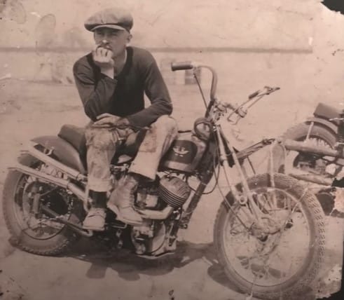 Kenny Von Dutch sits on his modified bobber motorcycle and seems to be deep in reflection resting his chin in his hand. Kenny was one of the pioneers of bobber custom builds in America.