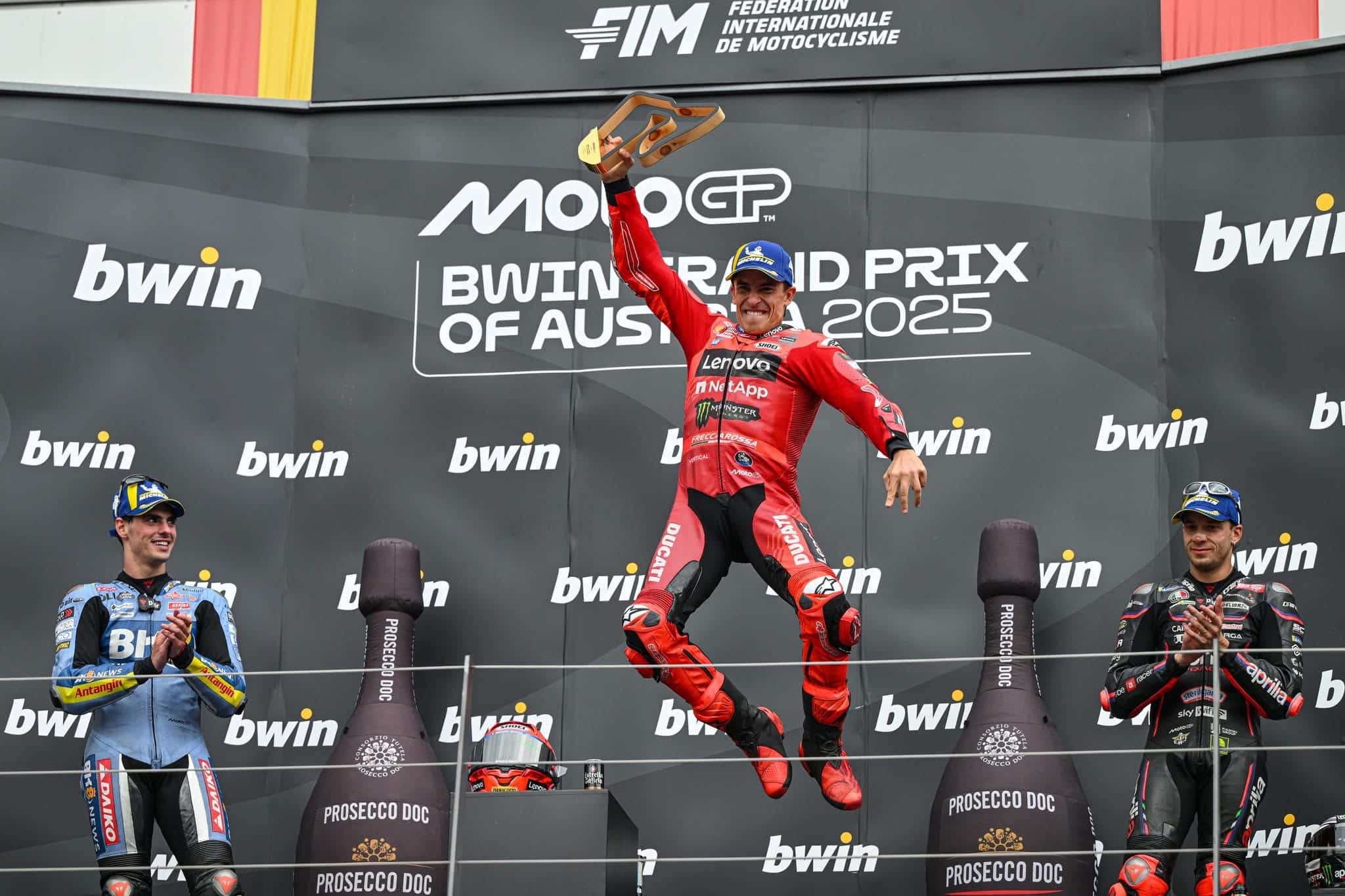 Marc Márquez jumps on the podium at the Austrian Grand Prix, trophy raised above his head, as second-place finisher Fermín Aldeguer and third-place Marco Bezzecchi celebrate alongside him