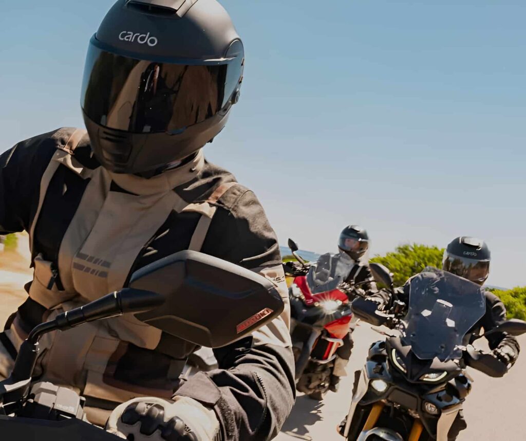 Cardo BEYOND GT and GTS First Look: Next-Gen Smart Helmets Three riders wearing Cardo BEYOND GT and GTS helmets showing integrated mesh intercom modules, 53 mm speakers, and aerodynamic shell design.