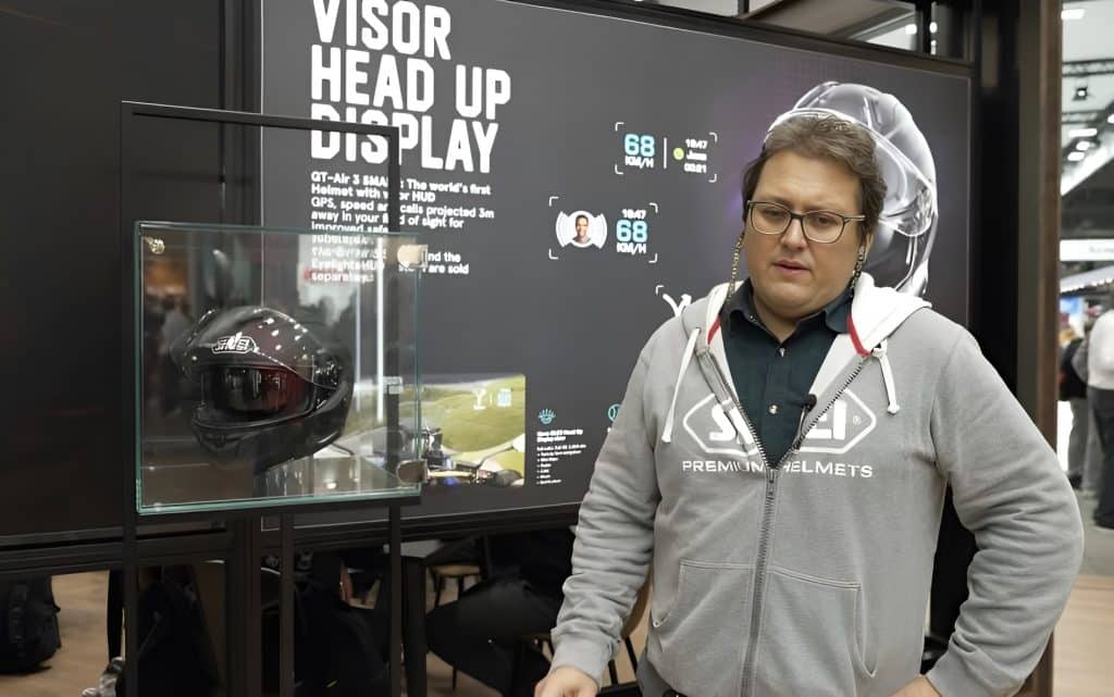 Shoei GT-Air 3 Smart First Look: AR-Integrated Helmet Fabian Naziruka stands next to a glass display case containing the the Shoei GT-Air 3 Smart helmet with EyeLights AR display. Behind him, a large screen reads "Visor Head Up Display" as he explains to me how the EyeLights HUD system will enhance rider safety and situational awareness by reducing glance-away time, delivering an intuitive low-distraction interface, and creating a scalable, connected platform that can add navigation, telematics and advanced alerts in future updates.